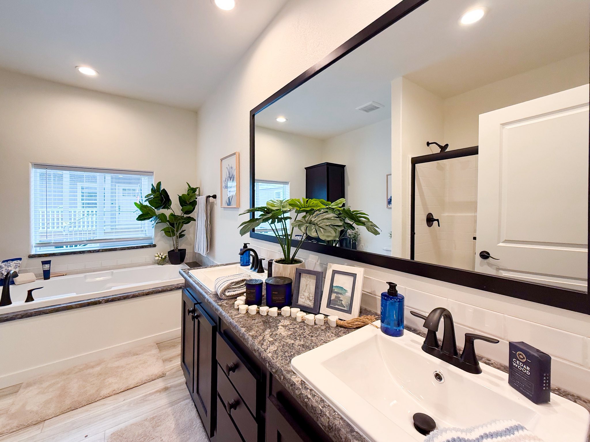 Modern bathroom with granite countertop, large mirror, and black fixtures. A plant and candles add a serene touch. Bathtub and window in the background.