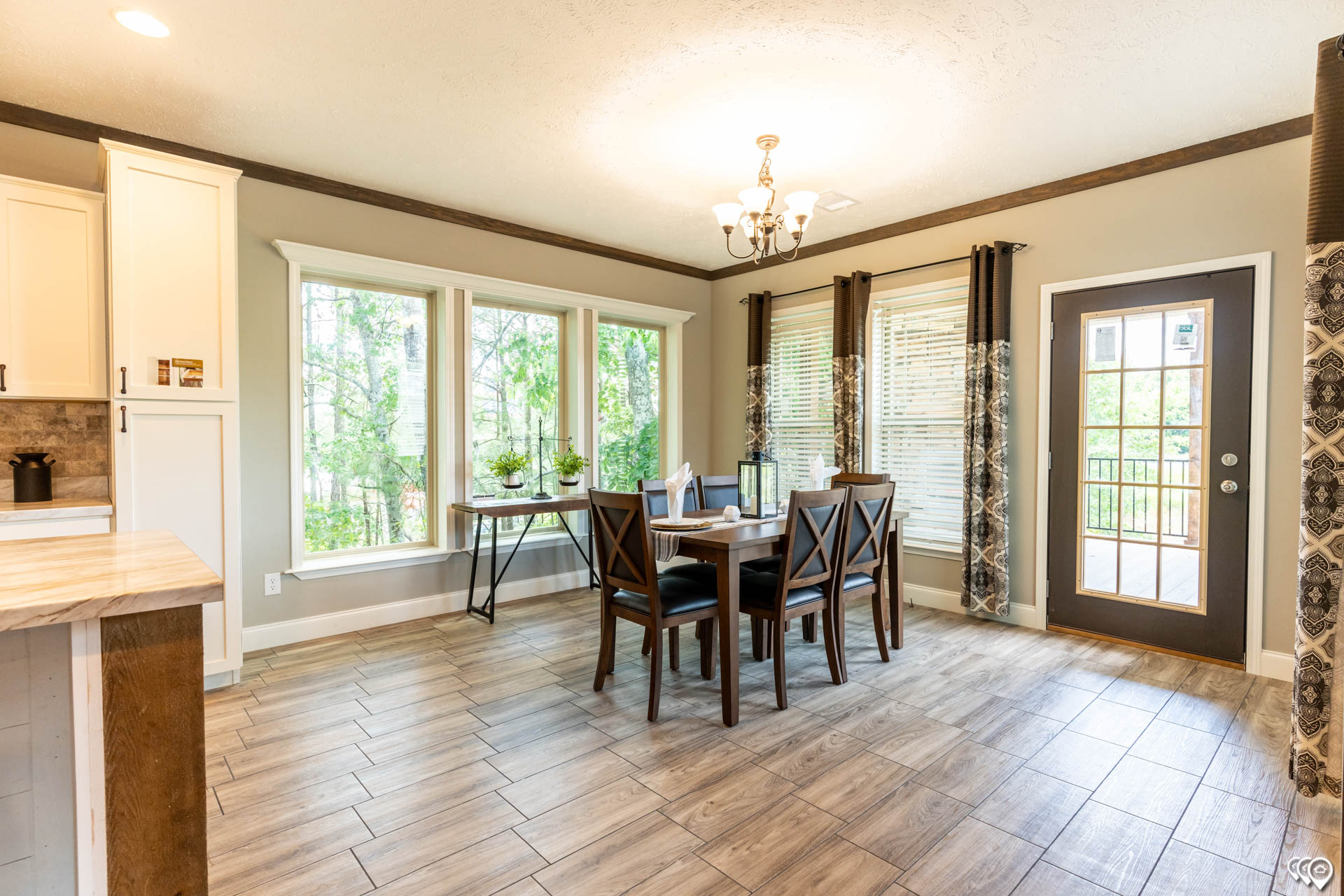 Bright dining area with a wooden table and four chairs under a chandelier. Large windows and a glass door offer views of greenery, creating an inviting atmosphere.