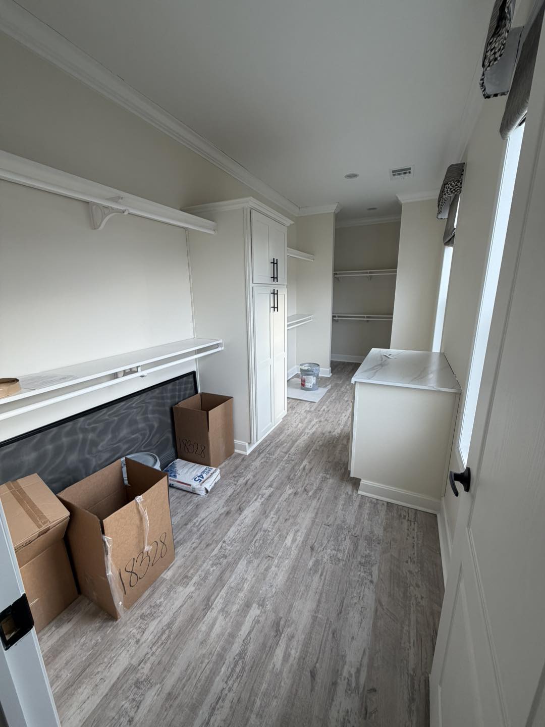 A narrow, empty walk-in closet with light wood floors, white walls, and built-in shelves. Open boxes and a paint can are on the floor, suggesting recent moving or renovation.