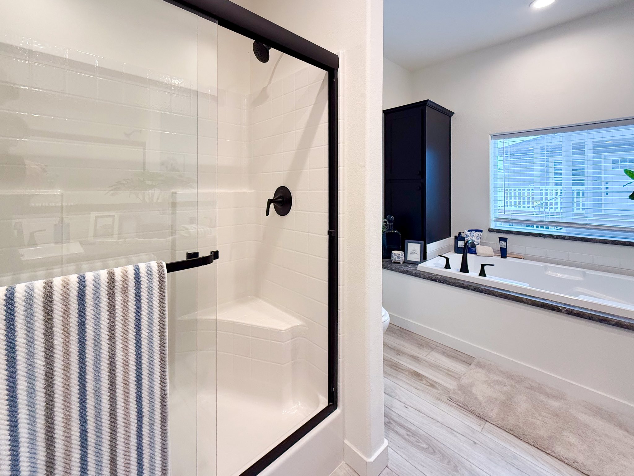 Modern bathroom with glass shower, black fixtures, and striped towel. Spacious bathtub by large window, light wood floor, and a cozy ambiance.