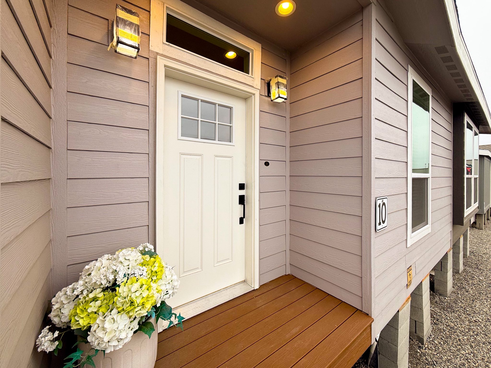 Front porch of a modern, light purple house with a white door and transom window. Potted white and yellow flowers sit by the entrance, under lit sconces.