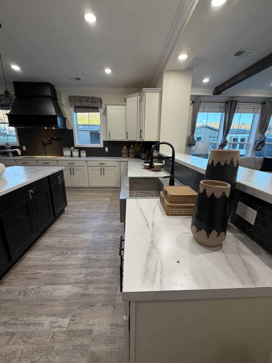 Modern kitchen with white cabinets, a black range hood, and gray flooring. Marble countertops feature decorative vases, giving a sleek, contemporary feel.