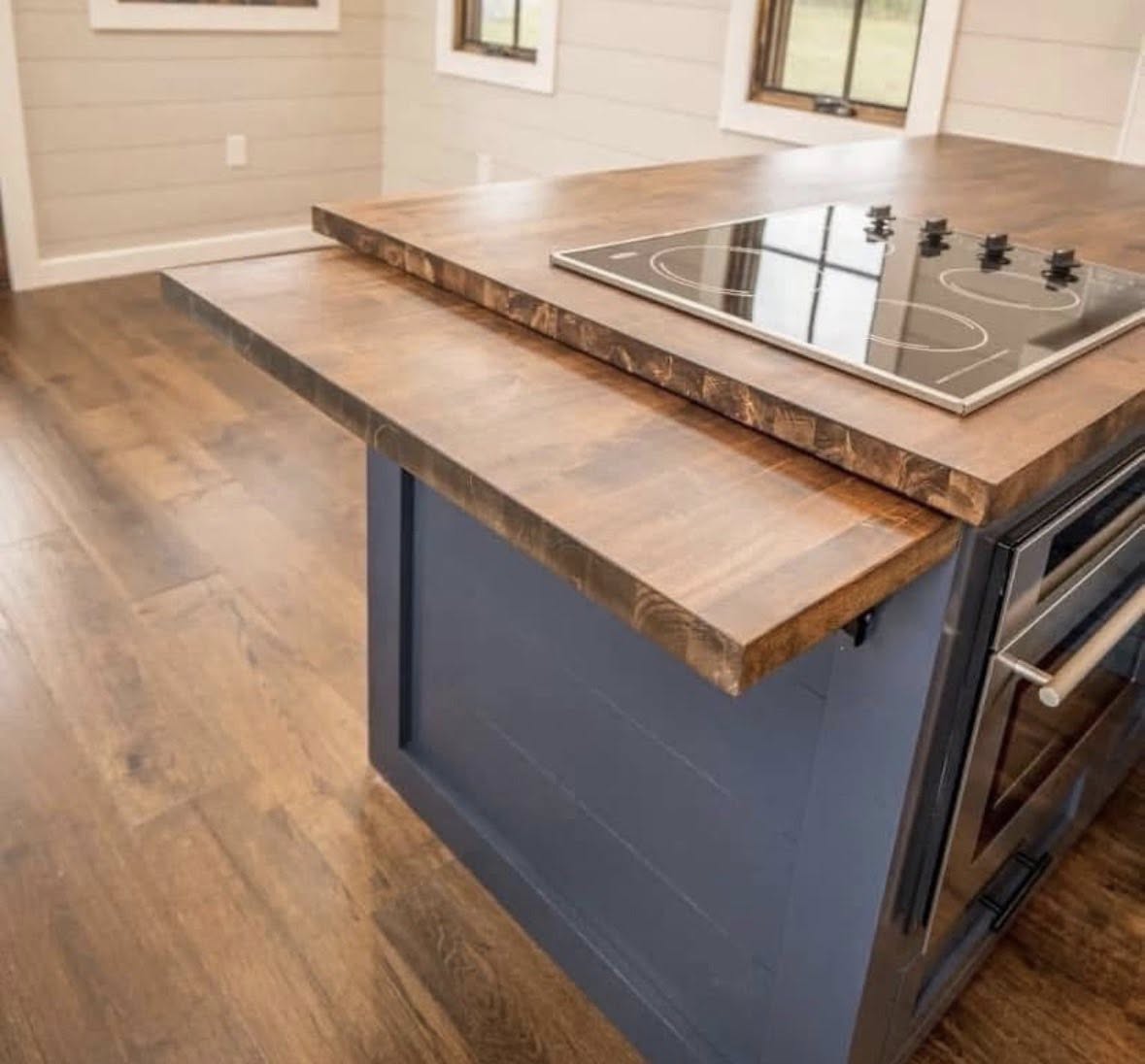 Rustic kitchen with wooden floor, featuring a blue island with a thick wooden countertop. A black glass stovetop is embedded. Warm, inviting atmosphere.