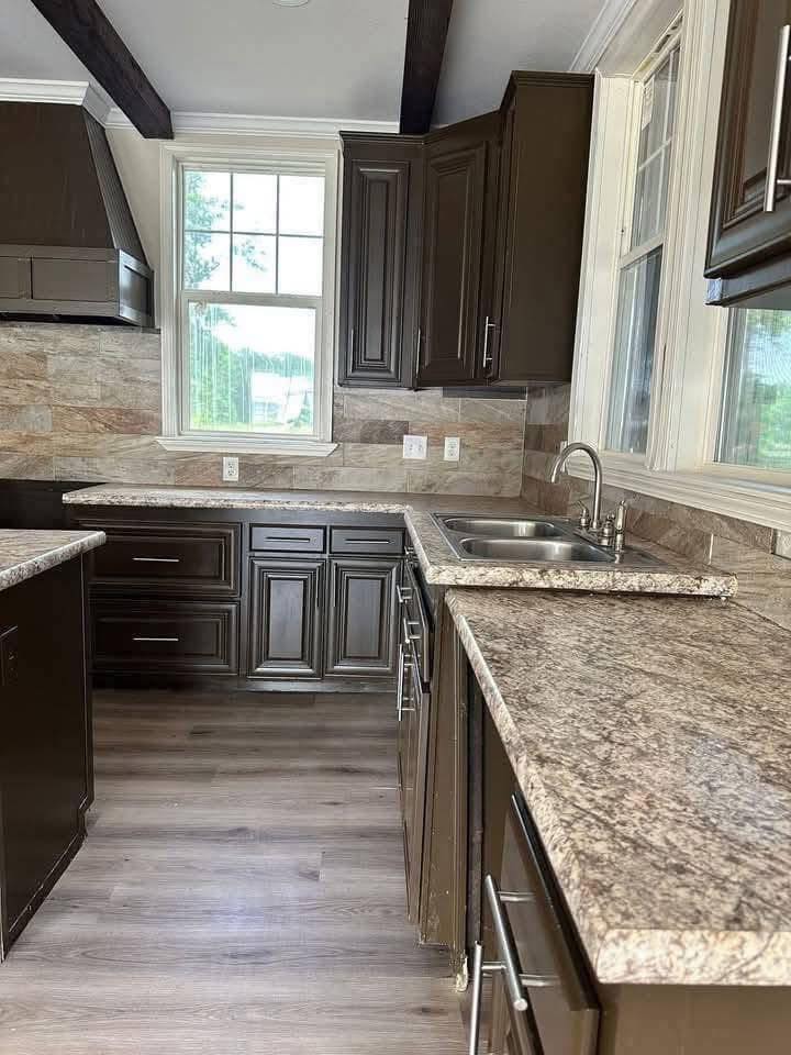 A modern kitchen with dark wooden cabinets and marble countertops under highlighted lighting. Hardwood floors and a window create a bright, inviting tone.