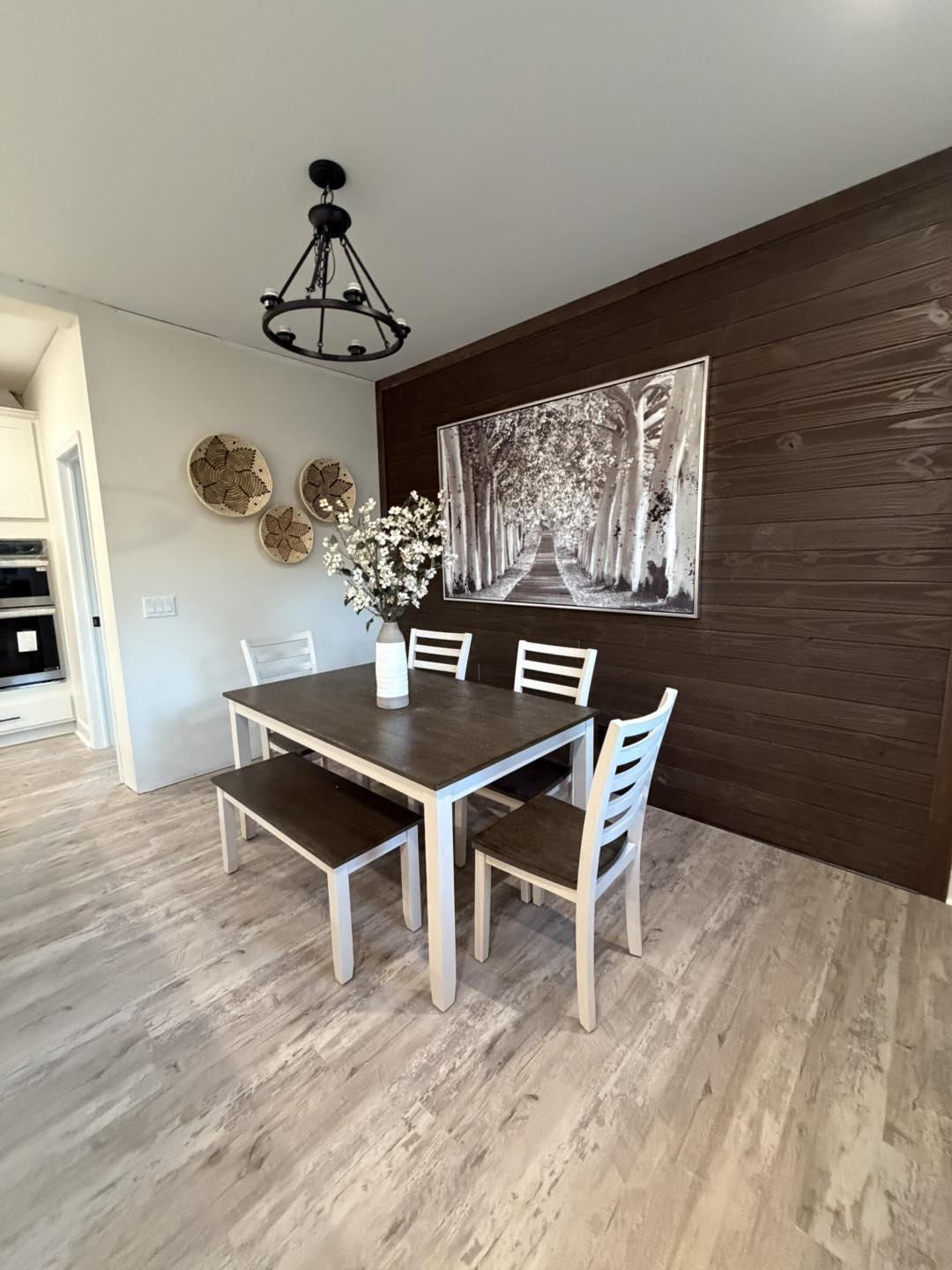 Dining area with wooden table, two chairs, and a bench. Dark wooden accent wall with a large black-and-white tree path photo. Light wood flooring, floral vase centerpiece, round wicker wall decor, and black metal chandelier enhance the rustic ambiance.