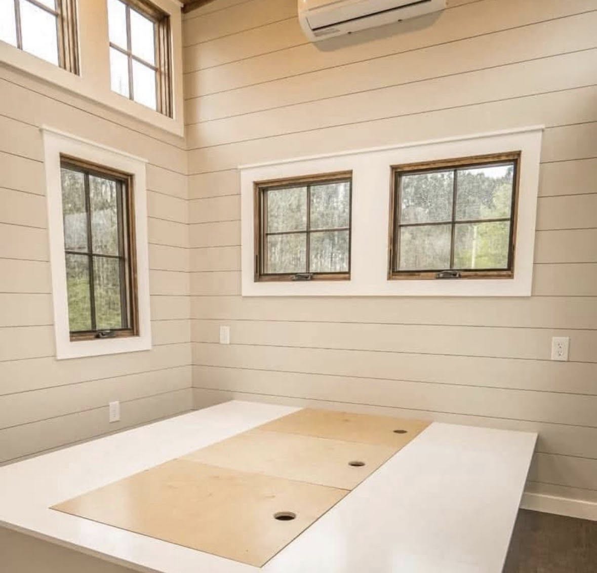 Bright room with beige shiplap walls, three windows with wooden frames, and a white ceiling. Air conditioner above. A large white surface with cutouts. Cozy and modern.