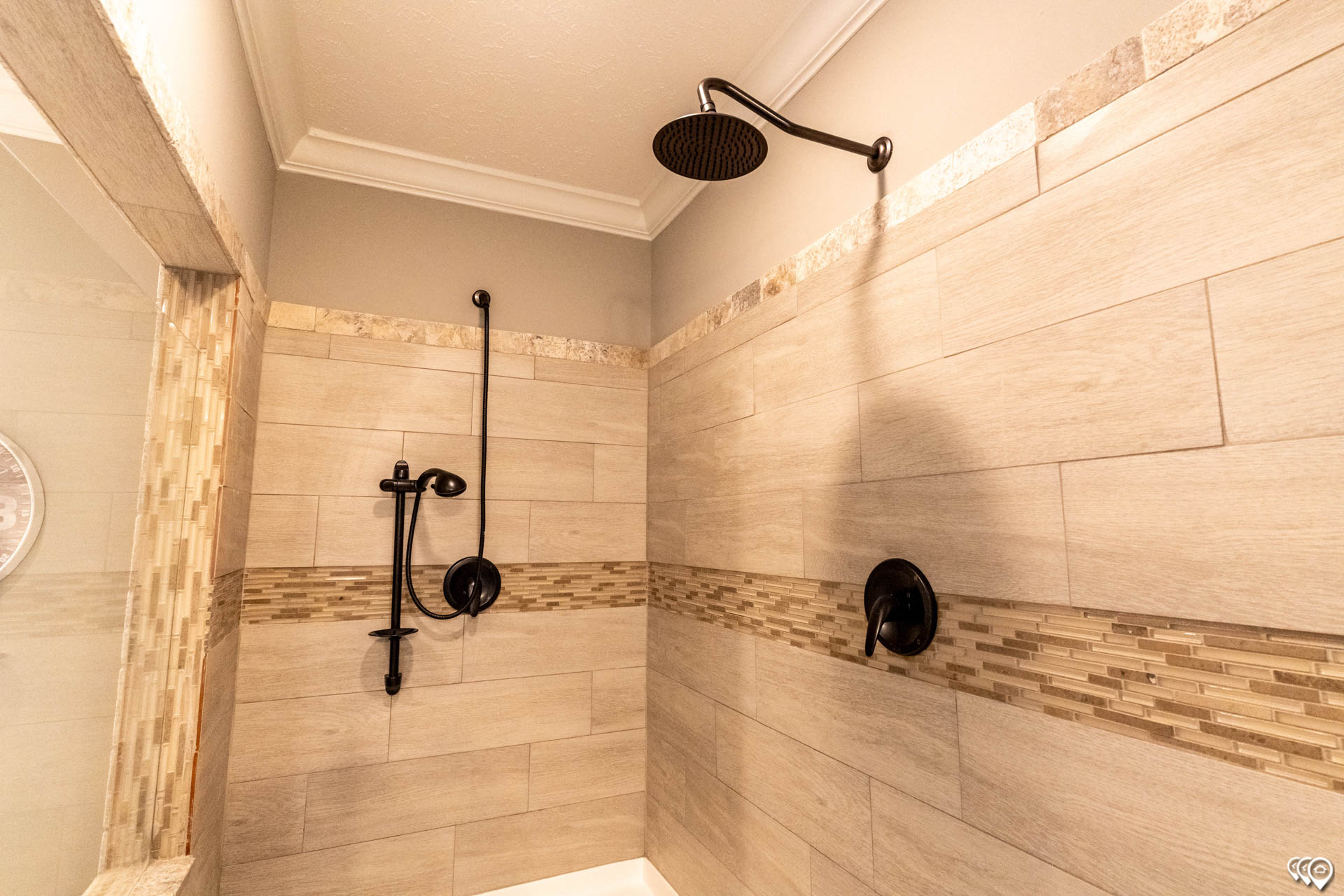 Modern shower with beige tiles and decorative mosaic strip. Features a black rain showerhead and handheld sprayer, creating a sleek, elegant look.