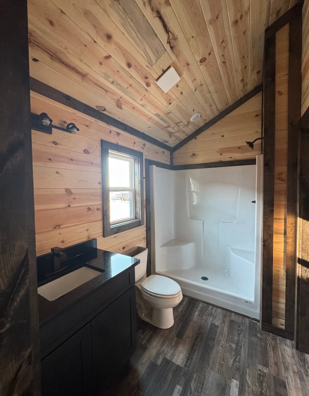 Bathroom with wooden walls and ceiling, featuring a window, white shower, and toilet. Dark vanity with a black countertop. Rustic and cozy ambiance.