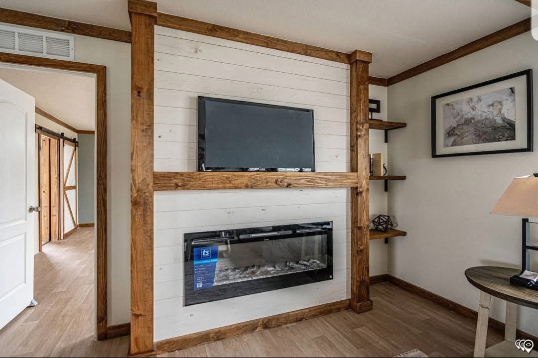 A cozy living room with wooden trim features a wall-mounted TV and a sleek electric fireplace beneath. Shelves hold decor, a lamp, and artwork adds warmth.