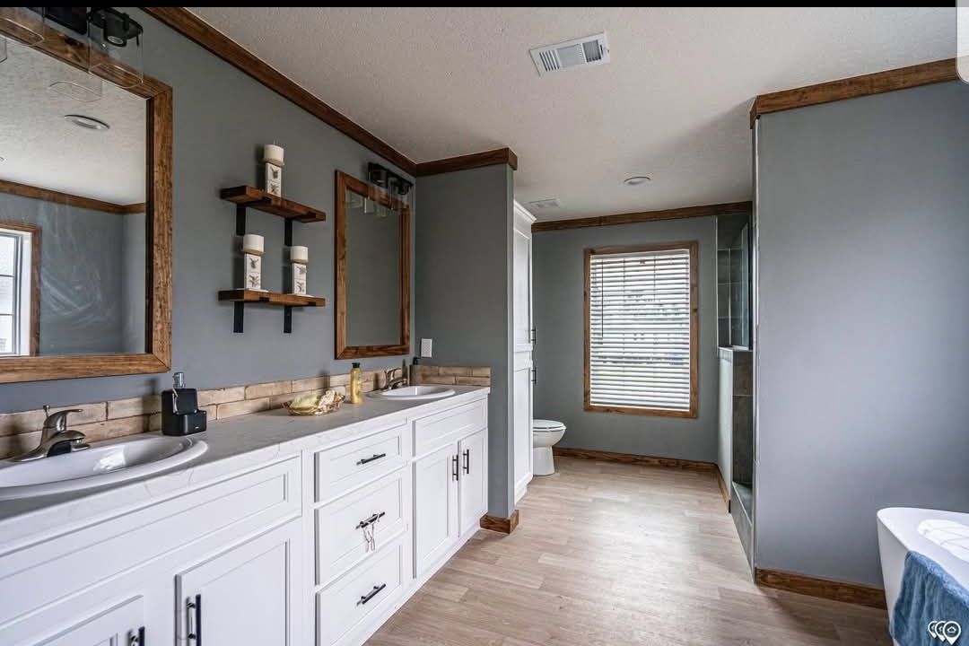 Spacious bathroom with gray walls and wood trim. Dual sinks with large mirrors, white cabinetry, and shelves. Window with blinds and adjacent shower.