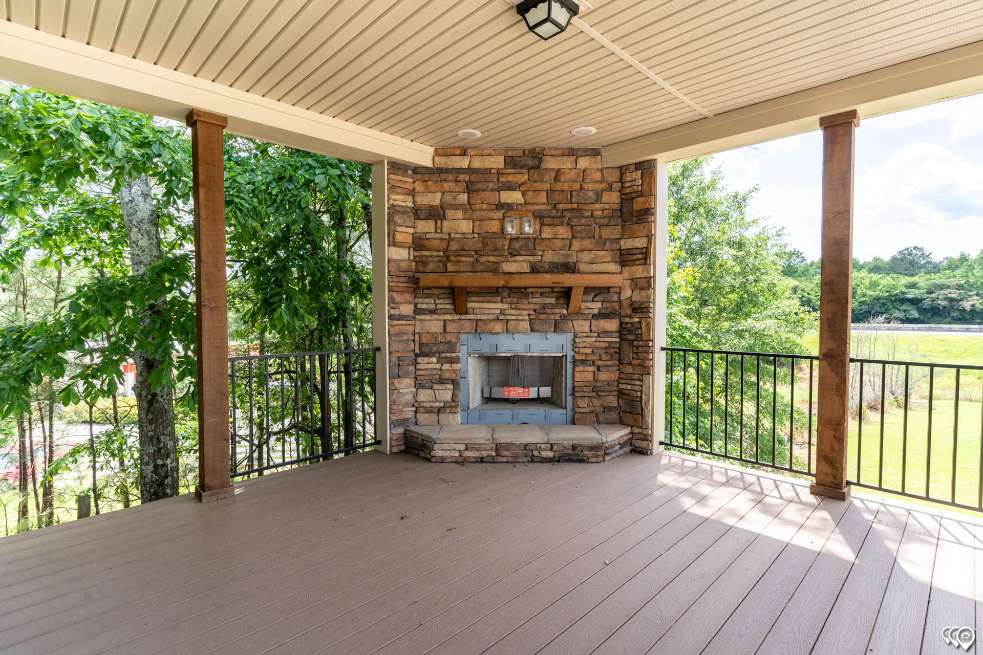 Covered patio with a stone fireplace, surrounded by trees and greenery. Features wooden floors, ceiling lights, and a serene view of a sunny landscape.