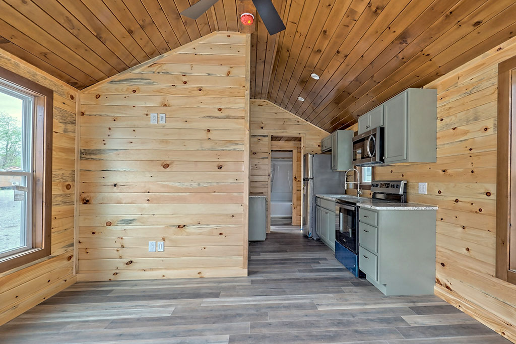 Cozy wooden cabin interior with light pine walls and ceiling. An open kitchen with gray cabinets and stainless appliances lines the right wall. A large window to the left brings natural light, highlighting dark wood floors.