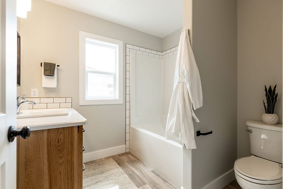 Modern bathroom with light beige walls and wood flooring. Features a white bathtub with subway tile, a wooden vanity, a toilet with a plant, and a white robe hanging, creating a serene and minimalist atmosphere.