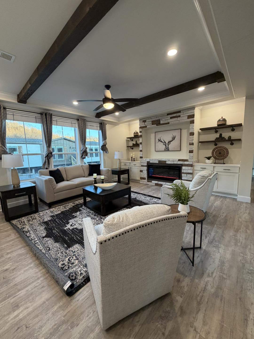 Modern living room with neutral tones. Features a beige sofa, patterned rug, fireplace with deer art, wooden beams, and large windows enhancing warmth.