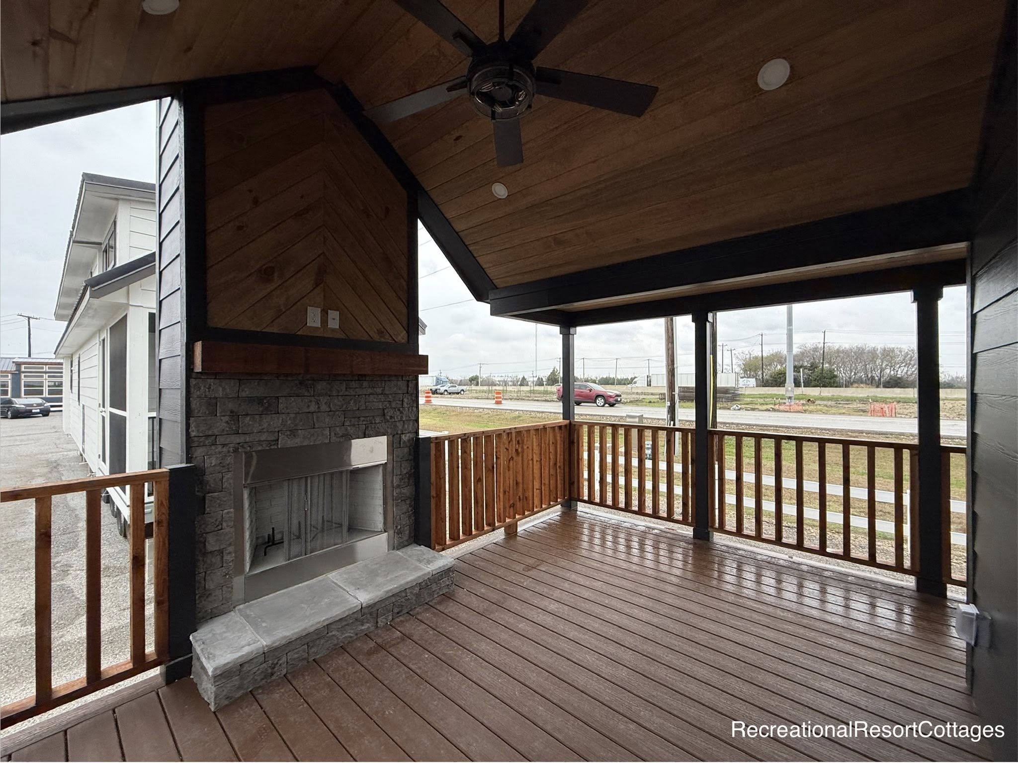 Covered patio with wooden floors, ceiling fan, and stone fireplace. Surrounded by a wooden railing, it overlooks a grassy area and road, creating a cozy and rustic atmosphere.