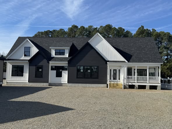 A modern house with a black and white exterior features a gabled roof and a spacious front porch. The setting is bright, with a clear sky and trees in the background.