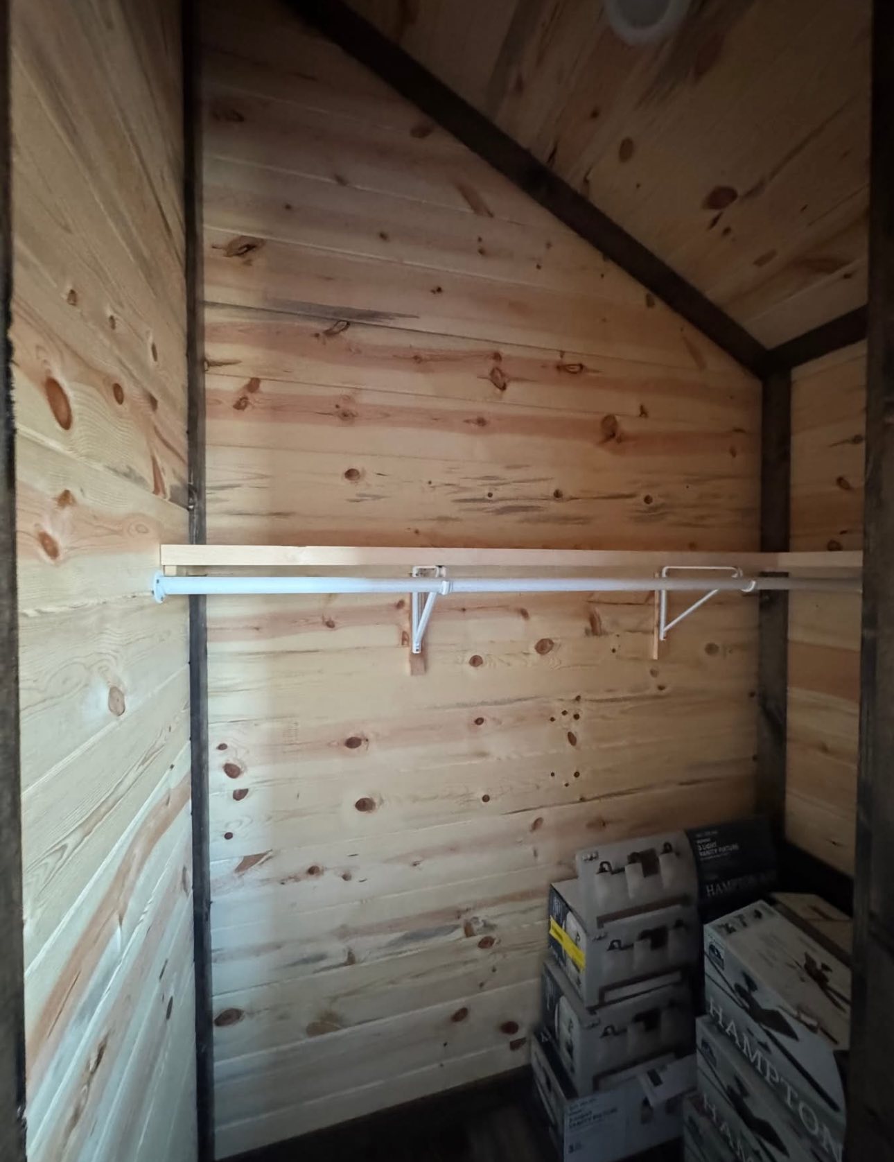 Wooden room with pine walls, featuring an empty white shelf supported by brackets. Several stacked boxes are in the corner, adding a rustic feel.