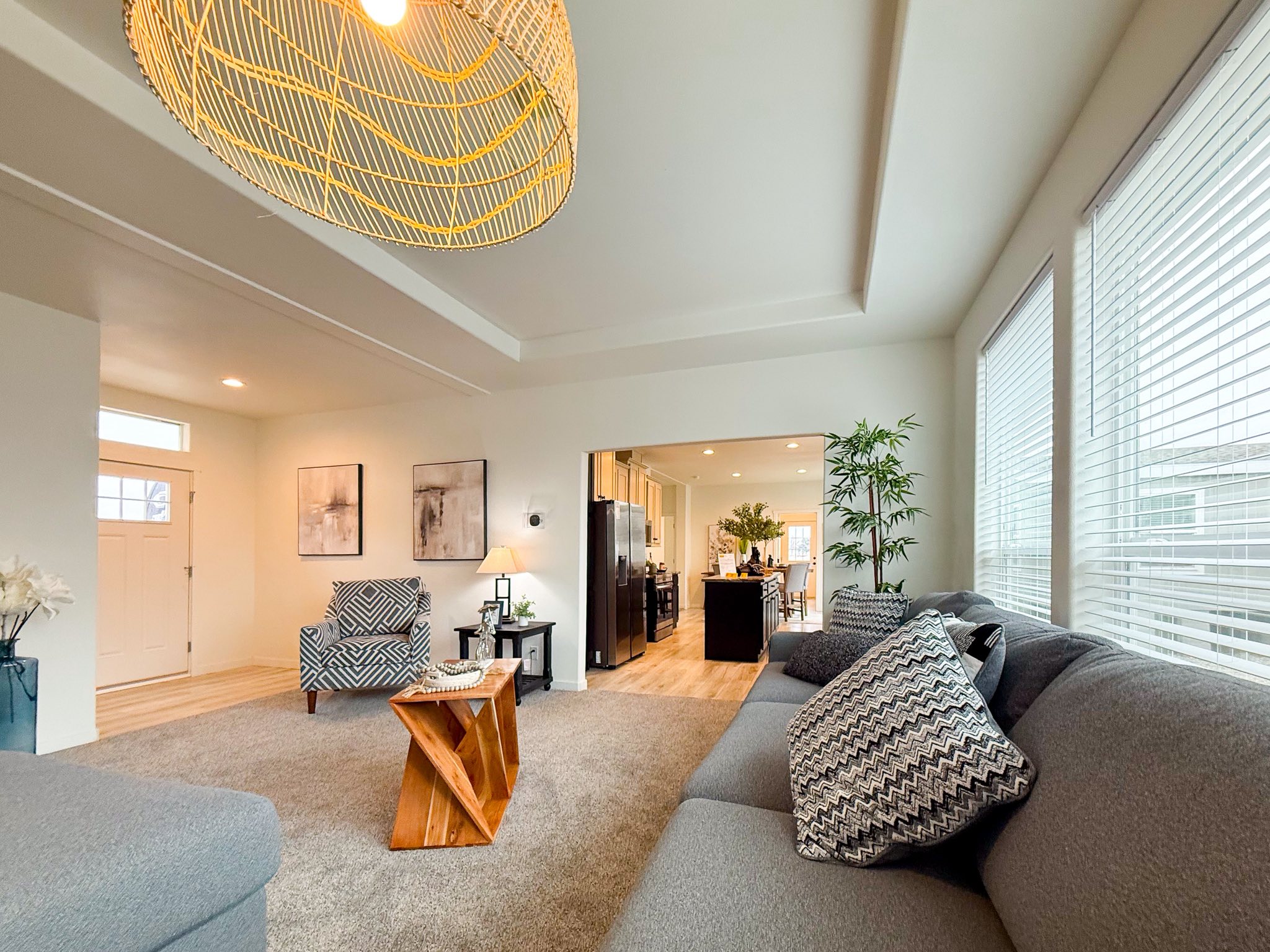 Spacious living room with gray sofa and patterned cushions, large windows, woven light fixture, and wooden coffee table. Open view to kitchen, warm ambiance.