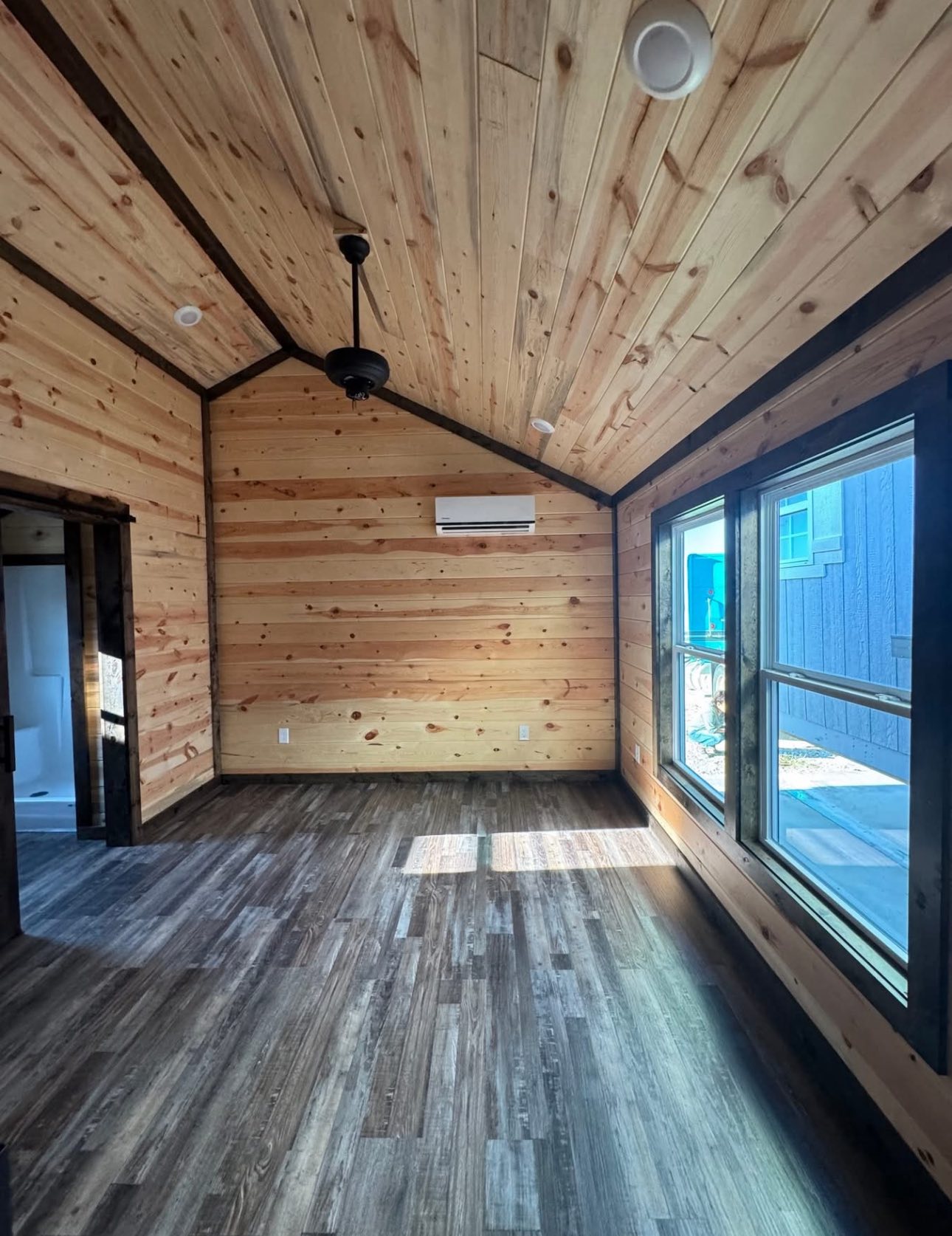 Cozy room with wood-paneled walls and ceiling, featuring a ceiling fan and large windows. Sunlight streams in, highlighting the dark wood floor.