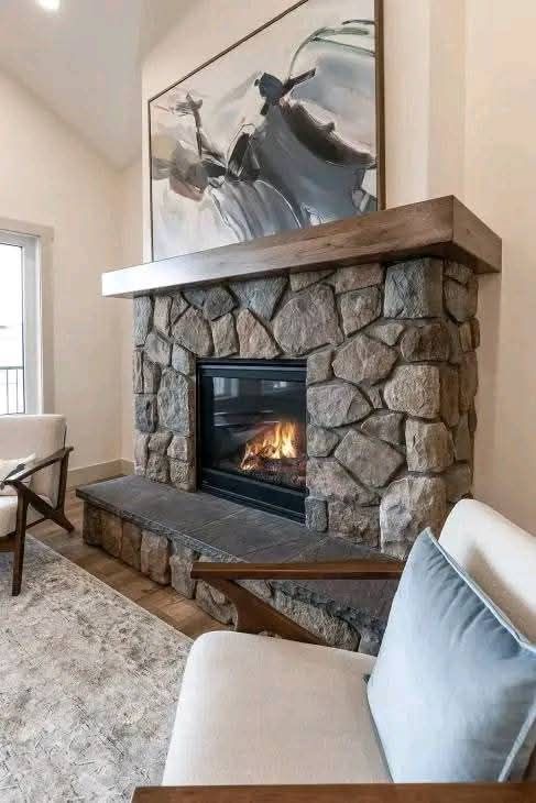 Cozy living room with a stone fireplace. A gentle fire burns below a modern abstract painting. Plush cream seating and a soft rug enhance relaxation.