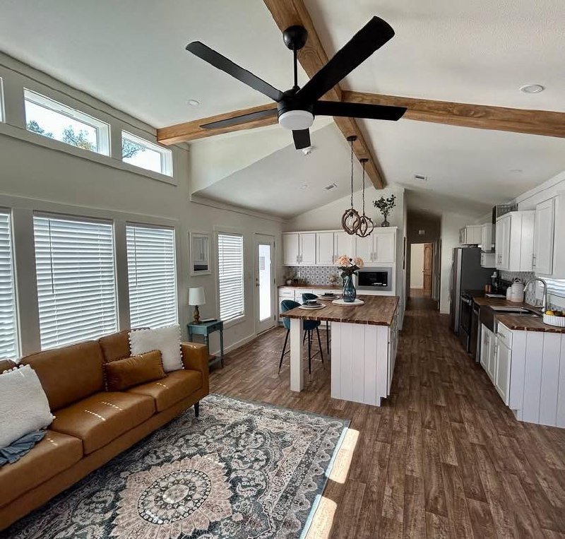 Modern living room and kitchen with vaulted ceiling, large windows, and wood beams. Features include a tan sofa, patterned rug, and island with stools.