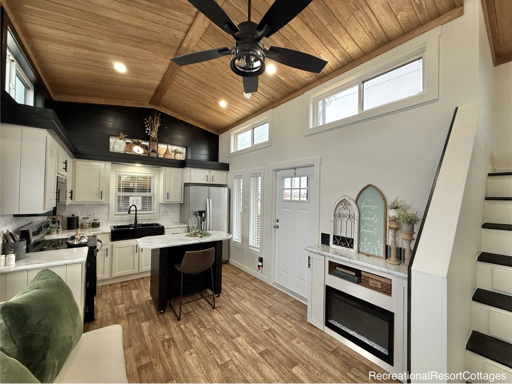 Cozy modern kitchen and living area with vaulted wood ceiling, black fan, white cabinets, central island, and decorative items on shelves. Warm, inviting ambiance.
