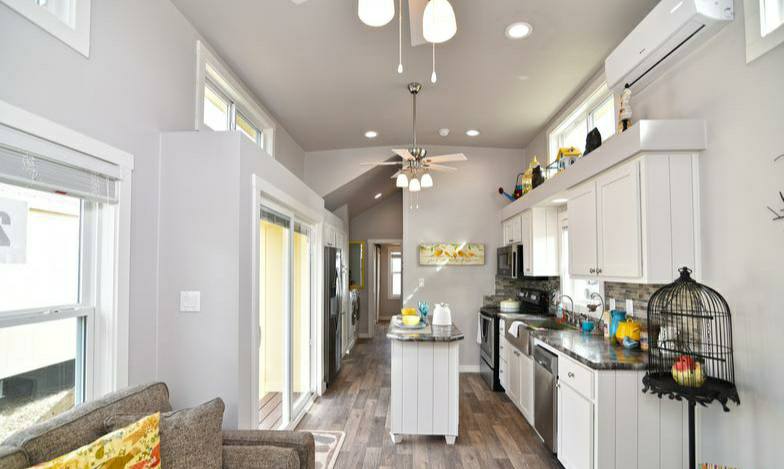 Bright, modern kitchen with white cabinets and stainless appliances. Central island, wooden floor, large windows, and a ceiling fan create an airy feel.
