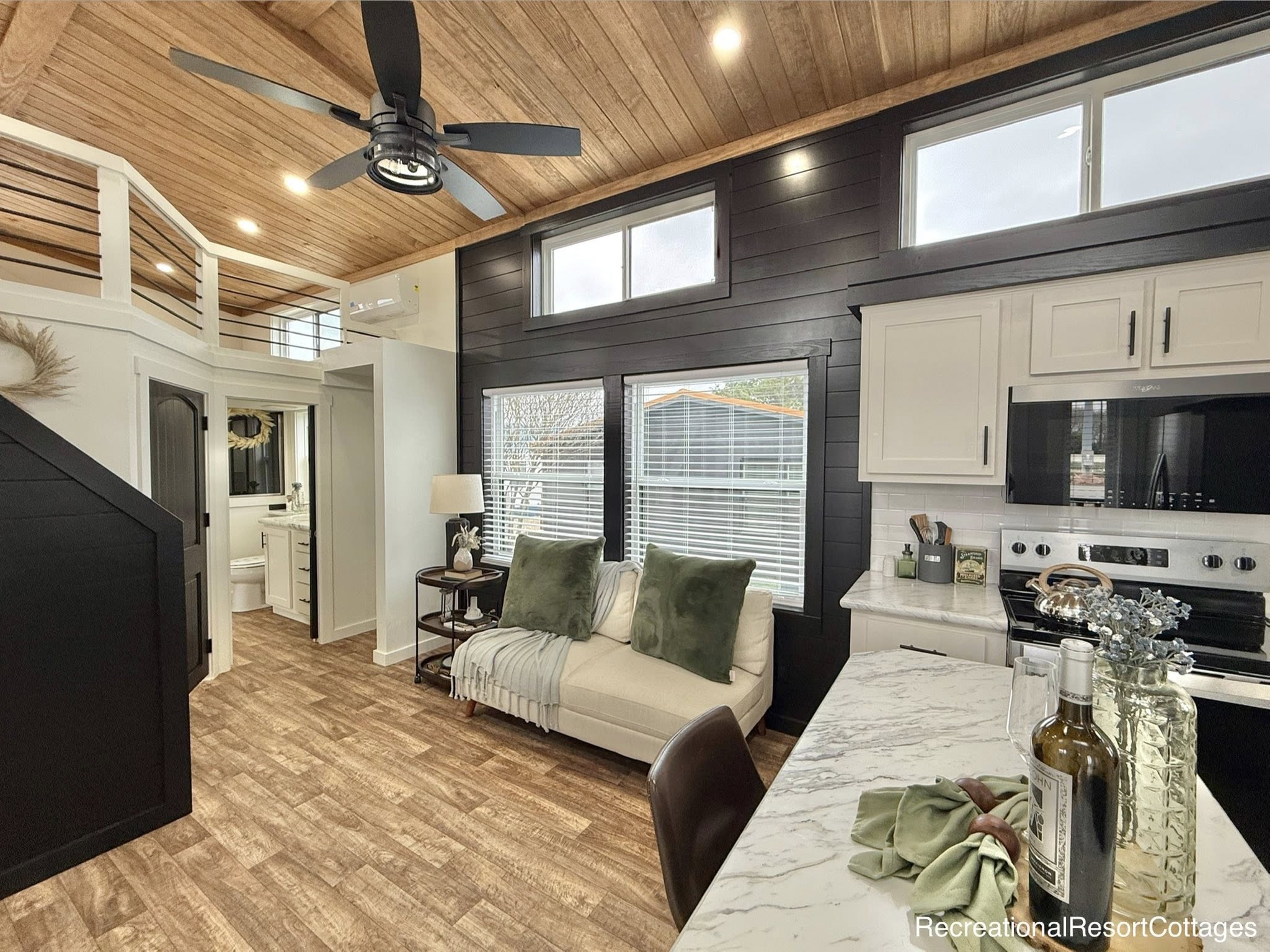Cozy open-plan tiny home interior with wood ceilings, black and white kitchen, a small loft, comfy couch with green pillows, and bright natural light.