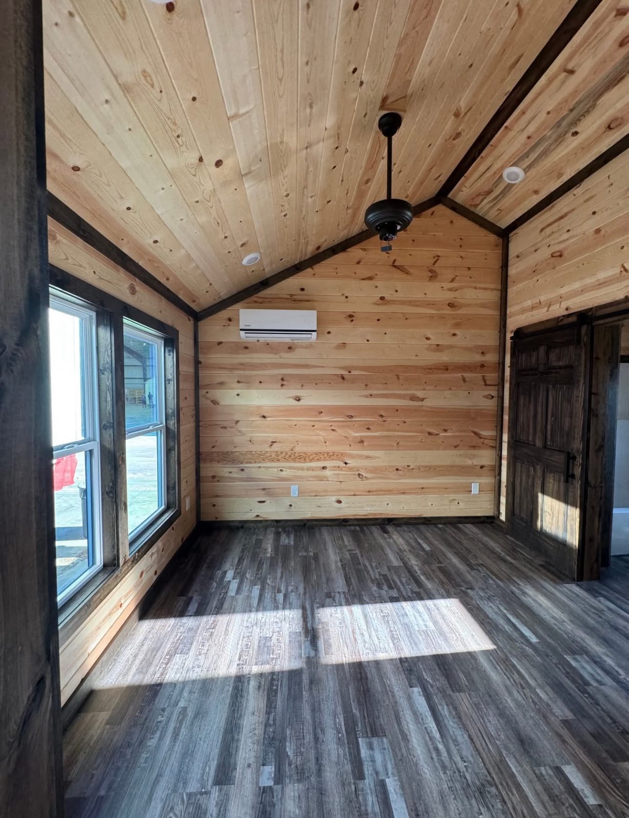 Cozy wooden room with sloped ceiling, large windows, and a ceiling fan. Light wood walls contrast with dark wood floors, creating a warm, rustic vibe.