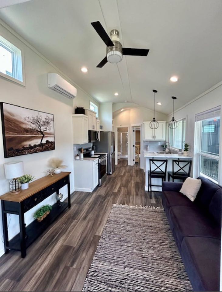 Modern tiny home interior featuring a sleek kitchen with white cabinets and stainless appliances. Cozy living area with black sofa, rug, and decor. Bright, airy ambiance.