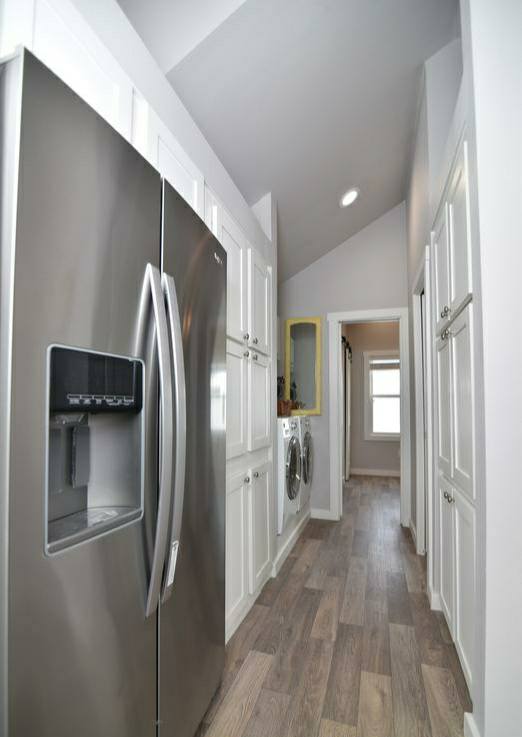 Narrow laundry room with gray flooring and white cabinets. A stainless steel fridge is on the left, beside a washer and dryer. Bright, modern feel.