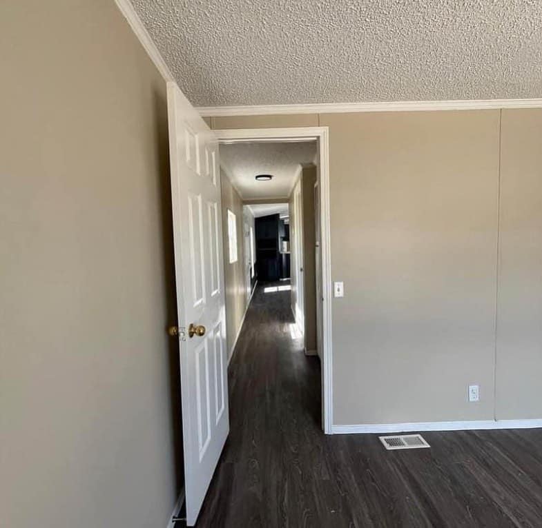 A hallway with beige walls and dark wooden flooring. A door is open, revealing a bright corridor leading to another room, creating a sense of depth.