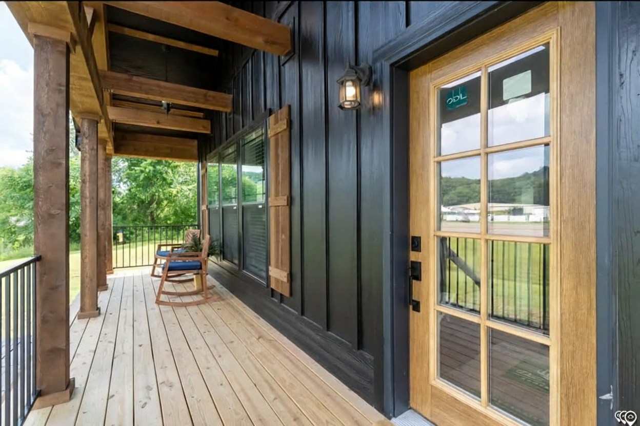 A rustic porch with a wooden floor and dark paneled walls, featuring a brown rocking chair. A glass-paneled wooden door is on the right, overlooking lush greenery.