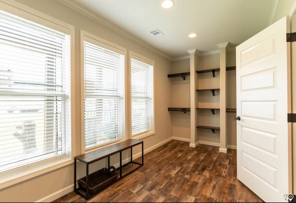 Bright room with wooden floor, beige walls, open door, and large windows with blinds on the left. Shelves on the back right wall add depth.