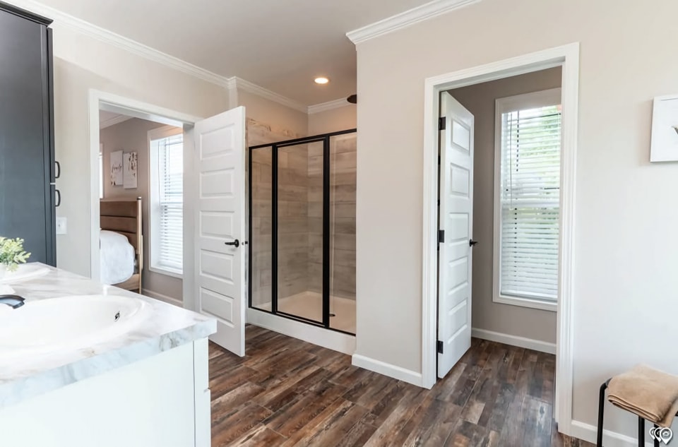 Modern bathroom with wood flooring and light walls. Features include a glass shower with black trim, marble countertop, and open doors to a bedroom.