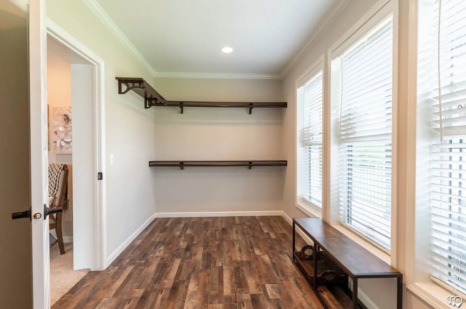 Bright, empty walk-in closet with wooden shelves on left wall, large windows on right with blinds, wooden floor, and small bench under windows.
