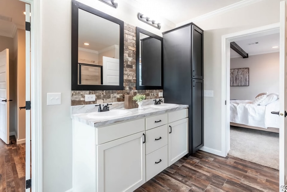 Modern bathroom with dual sinks, white cabinets, and a black cupboard. Two framed mirrors hang above. Adjacent bedroom with white bedding visible. Warm, cozy tone.