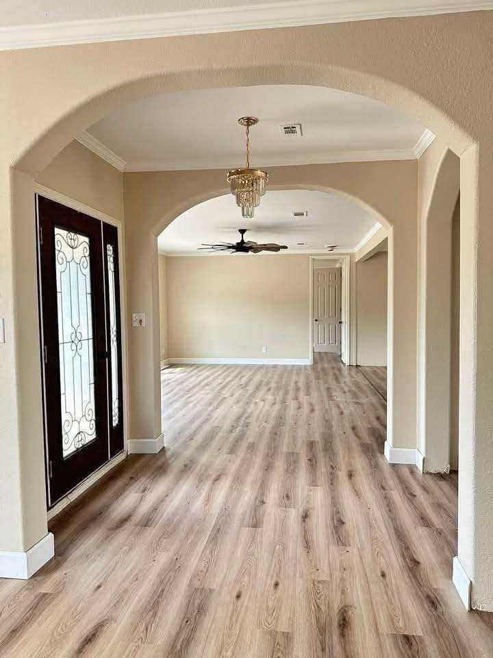 Wide hallway with light wood flooring, beige walls, arched entryways, decorative front door, chandelier, ceiling fan, and a closed door at the end. Cozy and elegant.