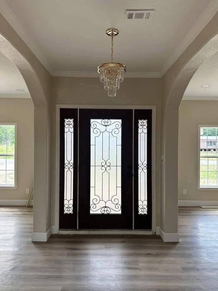 Elegant entryway with a dark wooden door featuring intricate glass designs. A chandelier hangs from the ceiling, and natural light streams in through side windows, creating a warm and inviting ambiance.