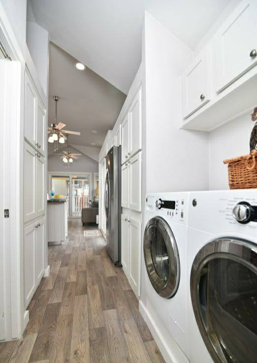 Bright laundry room with wooden floor, white cabinets, washer and dryer on the right. Hallway leads to a living area with ceiling fans. Cozy, organized ambiance.