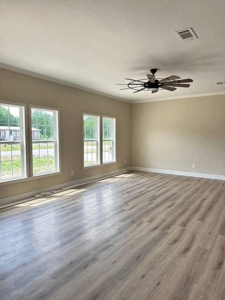Spacious, sunlit room with large windows, light wood flooring, beige walls, and a modern ceiling fan. The atmosphere feels open and airy.
