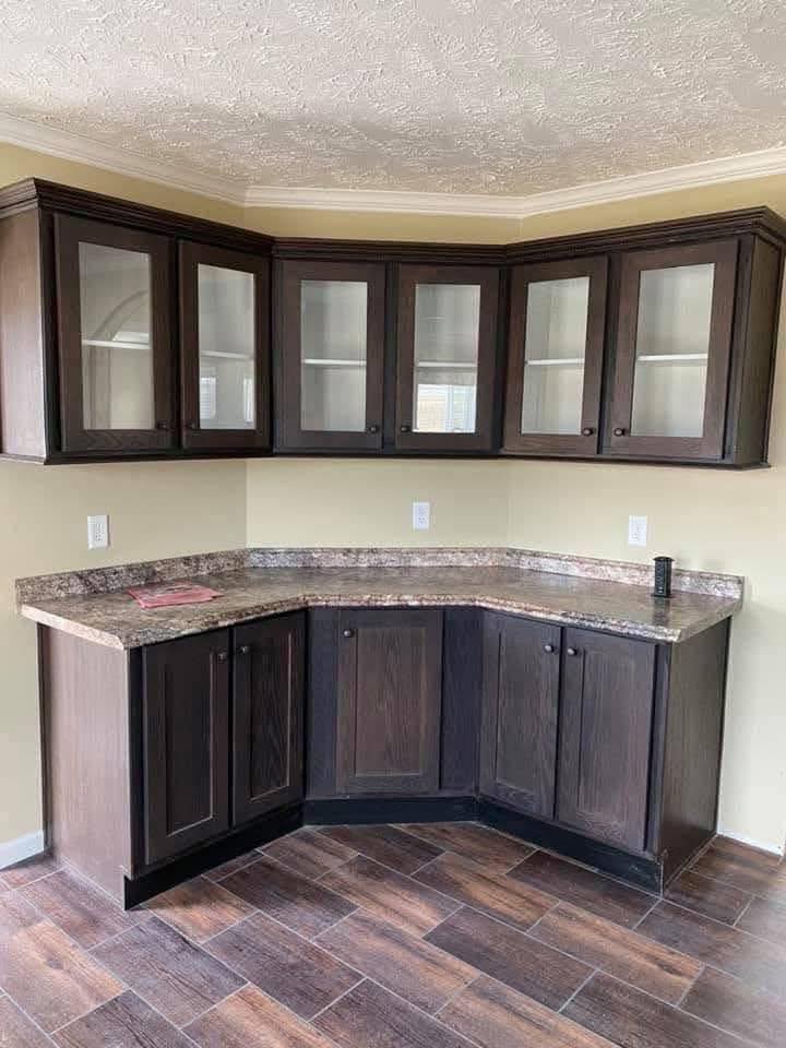 Corner kitchen cabinet setup with dark wood finish, glass-paneled upper doors, and marble-like countertop on brown tile flooring. Minimalistic and neat.