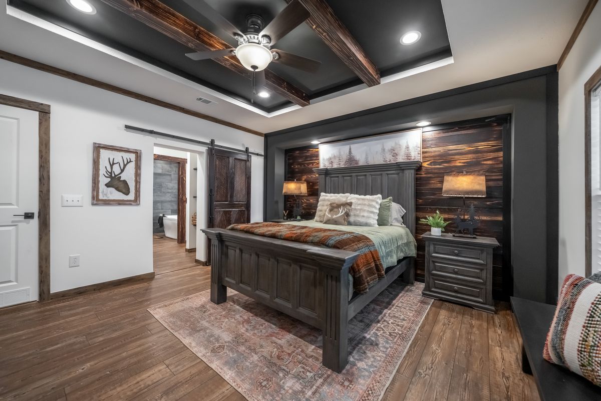 Rustic bedroom with wooden furniture, featuring a dark wood bed, plaid bedding, bedside lamps, and forest artwork. Warm lighting creates a cozy feel.