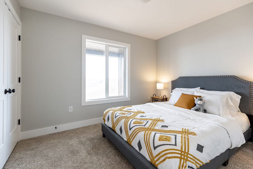A cozy bedroom with soft gray walls features a bed with a gray tufted headboard, white and gold-patterned bedding, and a small stuffed animal. A nightstand with a lit lamp adds warm light beside a window.