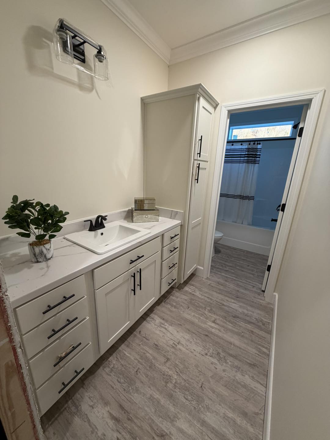 Modern bathroom with light wood flooring and white cabinetry. Features a sleek sink, black fixtures, a small plant, and a view into a shower area. Clean and elegant design.