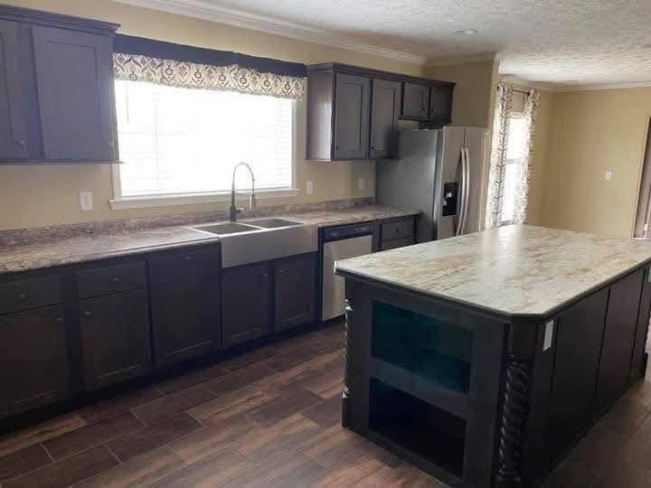 Spacious kitchen with dark wood cabinets, a large island, stainless steel appliances, and a farmhouse sink under a patterned valance. Warm and modern tone.
