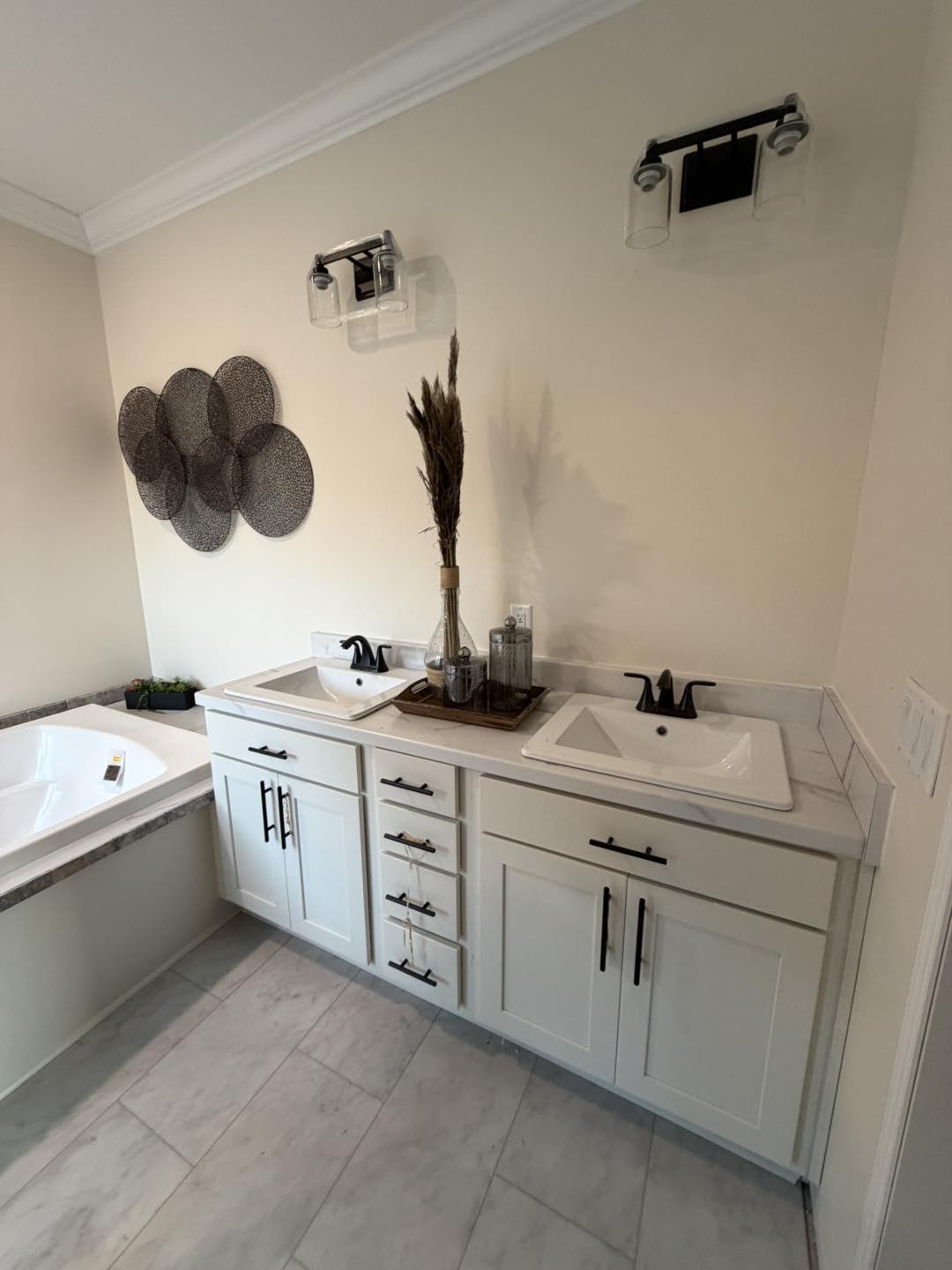 Modern bathroom with dual sinks, black fixtures, and white cabinets. Wall art features circular designs. Decorative tray with vases adds elegance.