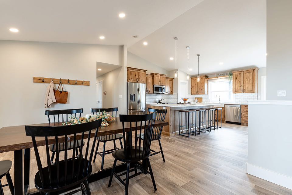 Spacious kitchen and dining area with wooden floors, black chairs, and a large table. Kitchen has wooden cabinets, stainless steel appliances, and bar seating. Cozy and inviting.