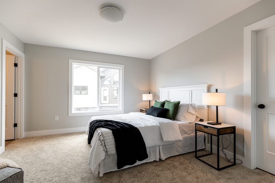 Bright bedroom with a neatly made bed featuring white sheets, green and black pillows, and a dark throw. Two bedside tables with lamps flank the bed, and a large window lets in natural light. Gray walls and beige carpet create a serene atmosphere.