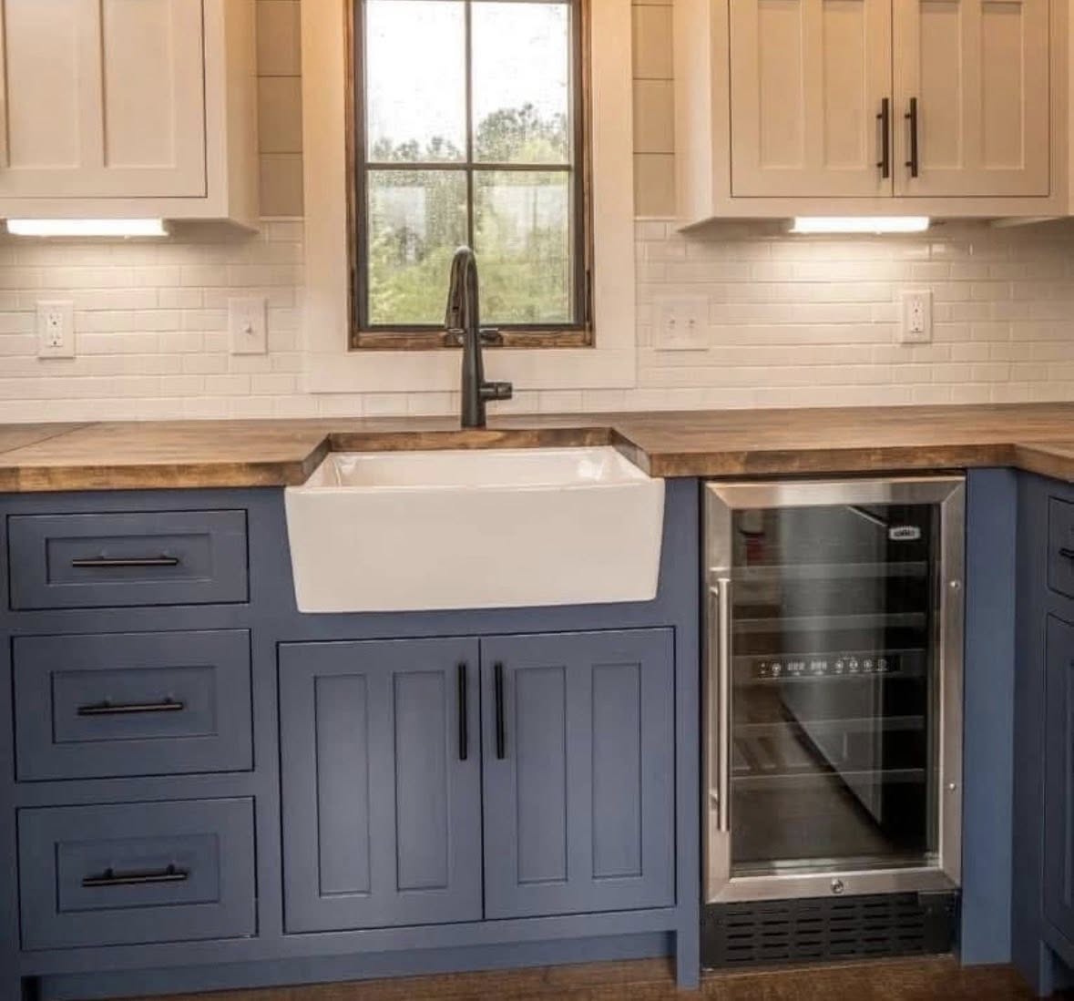 A cozy kitchen with blue cabinets, white farmhouse sink, wooden countertop, and stainless steel wine cooler. Warm lighting and a window add charm.